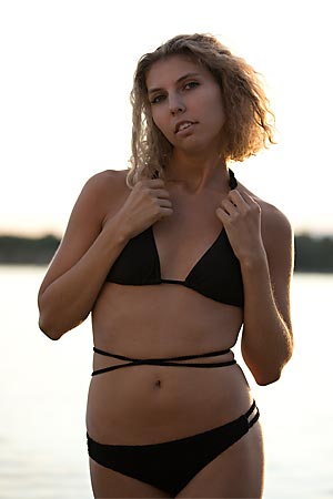 A young woman wearing swimwear is standing in front of a lake.