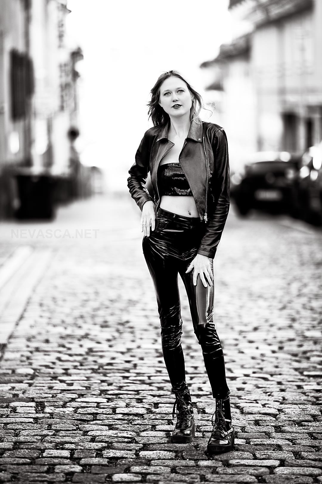 A young woman wearing leather trousers is standing on a cobble stone pavement.