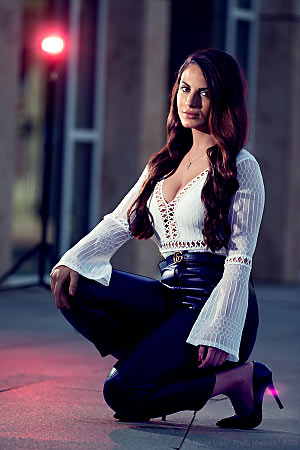 A young woman wearing leather trousers is kneeling in front of a building. In the background, there is a red light.