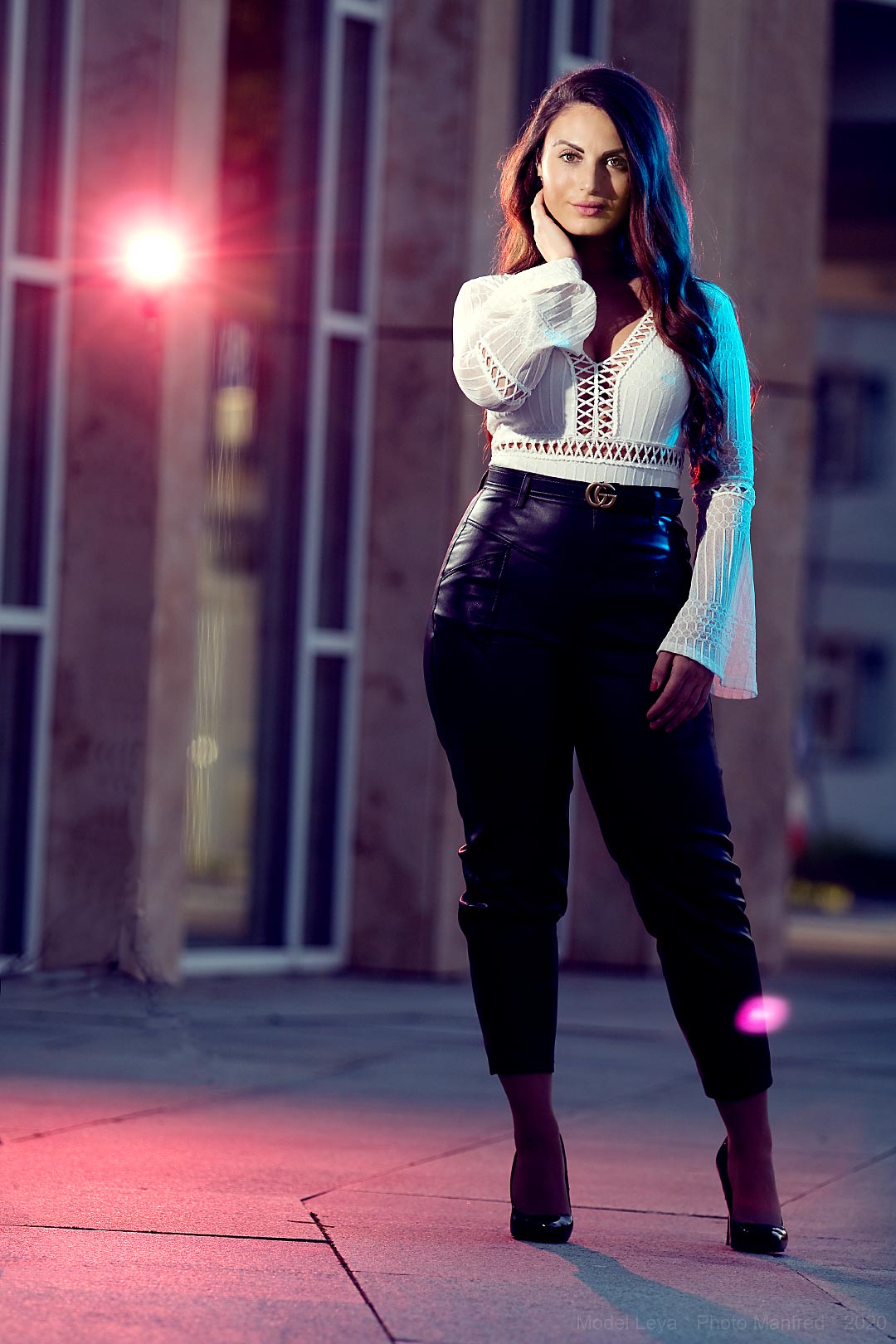 A young woman wearing leather trousers is standing in front of a building. In the background there is a red light.