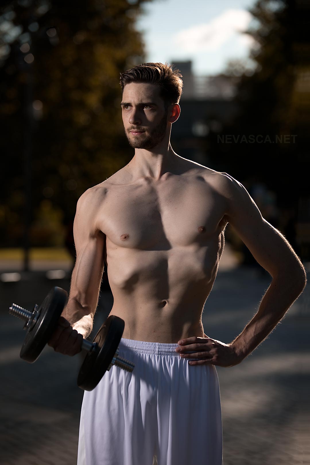 A young man standing in a park is holding a dumbbell.