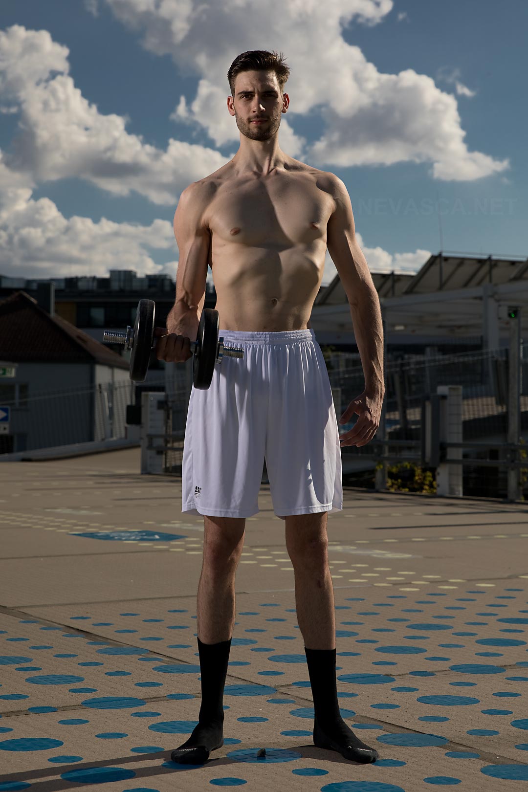 A young man standing on a parking space is holding a dumbbell.